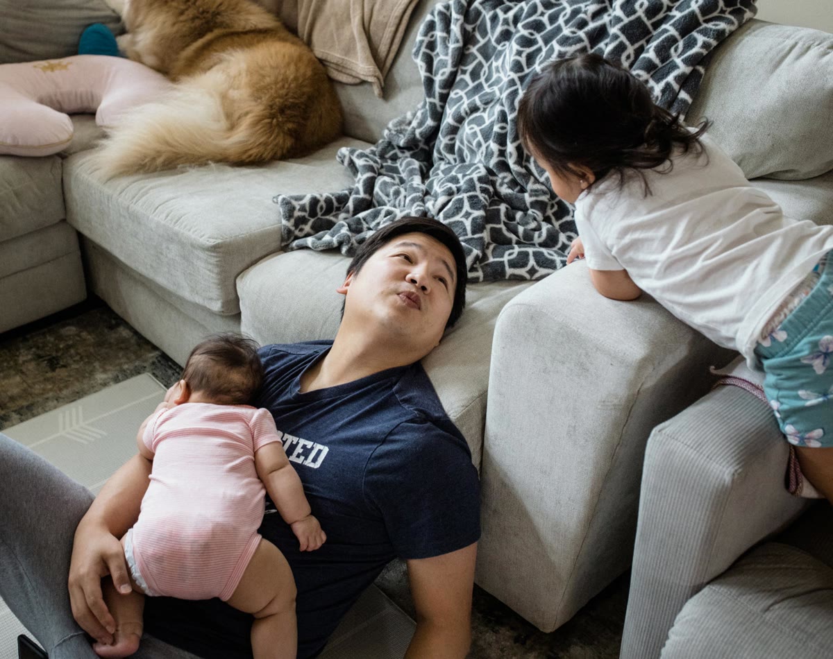A 35-year-old man holds his baby and talks to his toddler while sitting on the floor leaning against a couch.