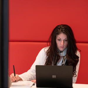 A woman writes in a notebook while looking at a laptop screen.