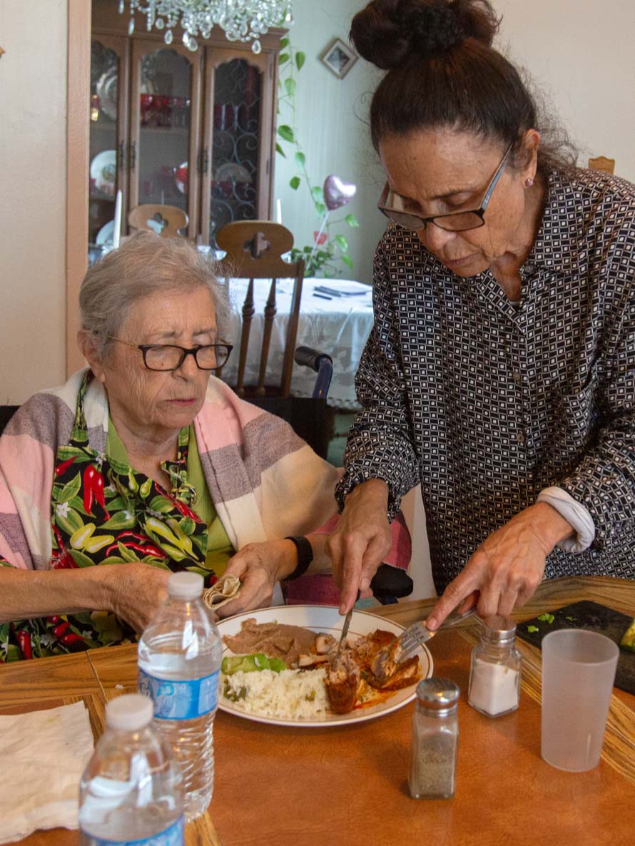 A woman stands using a fork and knife to cut food on the plate of an older person, who sits at a table.