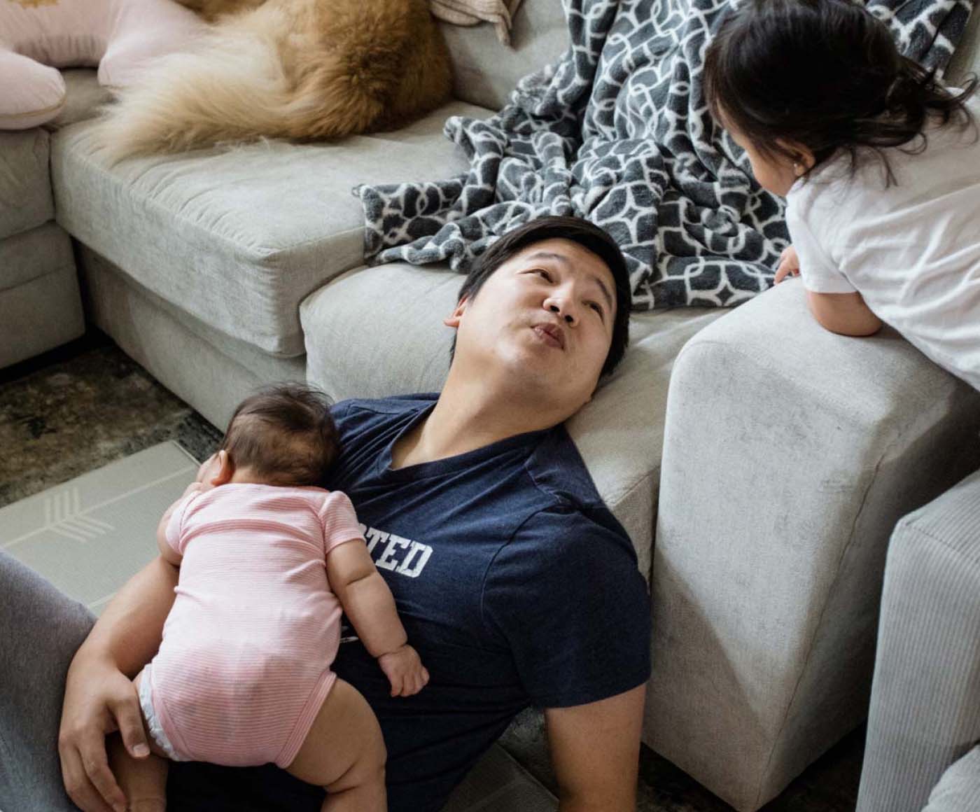 A father sitting on the floor of his living room talking to his child, with his baby in his lap