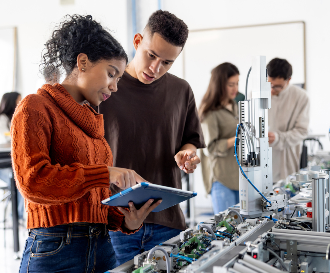 A woman shows a tablet screen to a peer in a robotics lab.