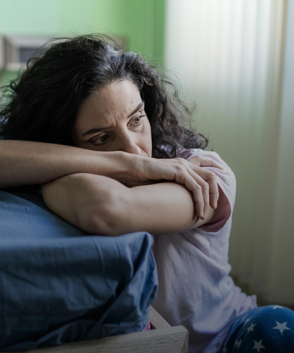 A worried woman sitting on the floor next to her bed.