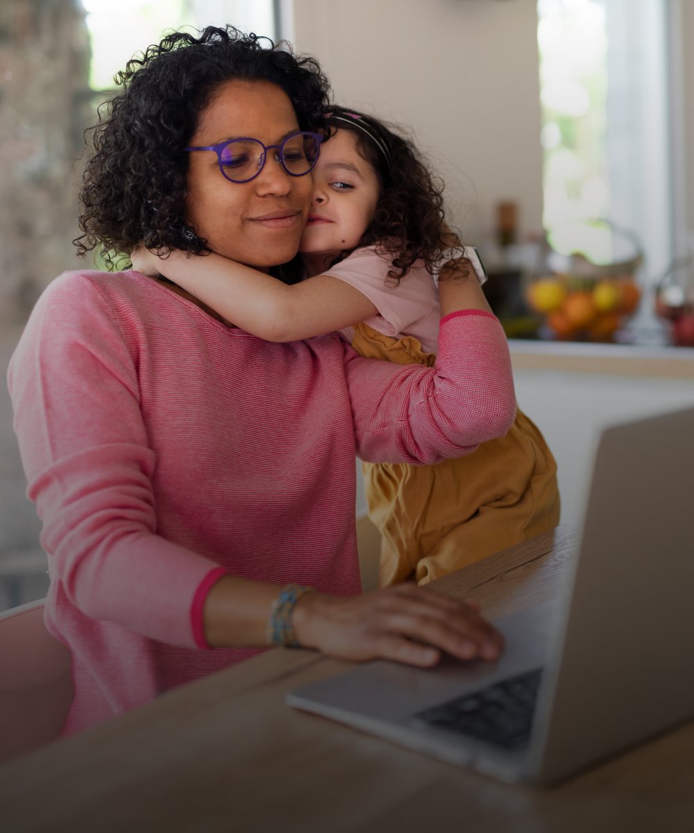 A mother hugging her daughter with one hand, and working on her computer with the other.