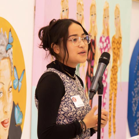 A person stands at a microphone in front of a wall decorated with art.