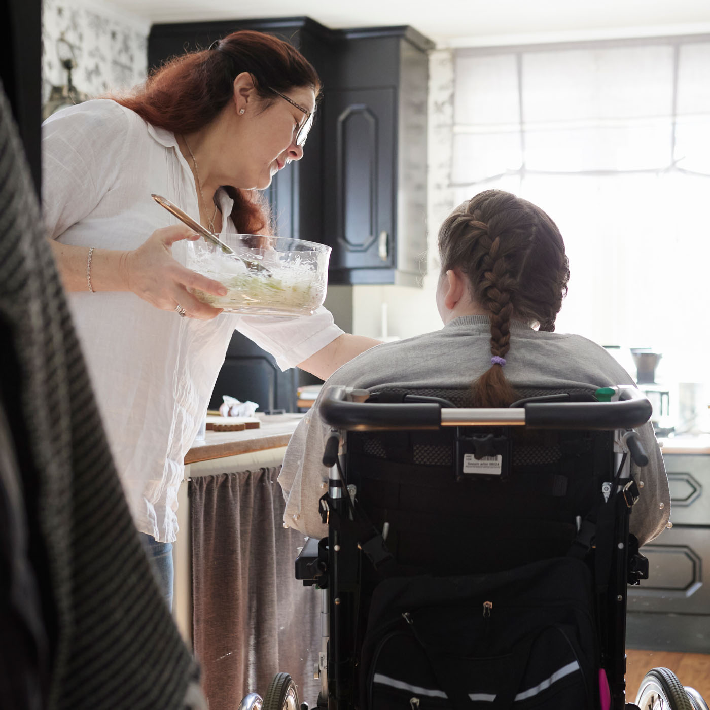 A mother feeding her daughter, who is in a wheelchair, while the father prepares food
