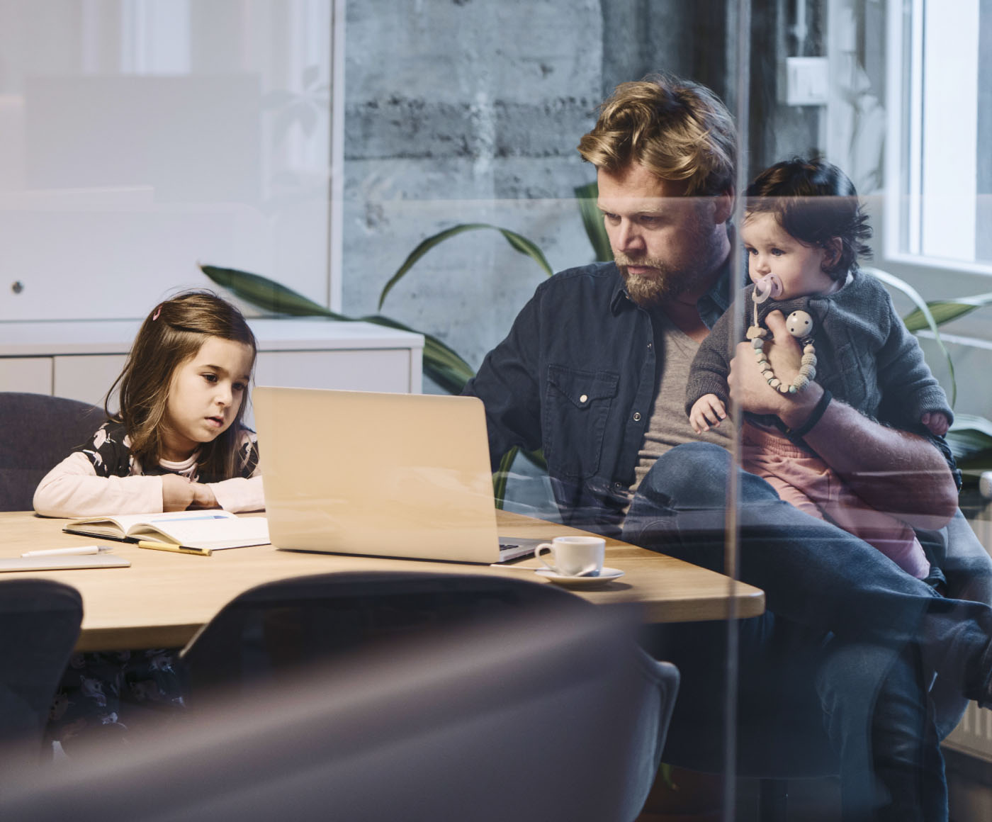 A man working on his laptop in the conference room of his office, with his two young daughters watching him work.