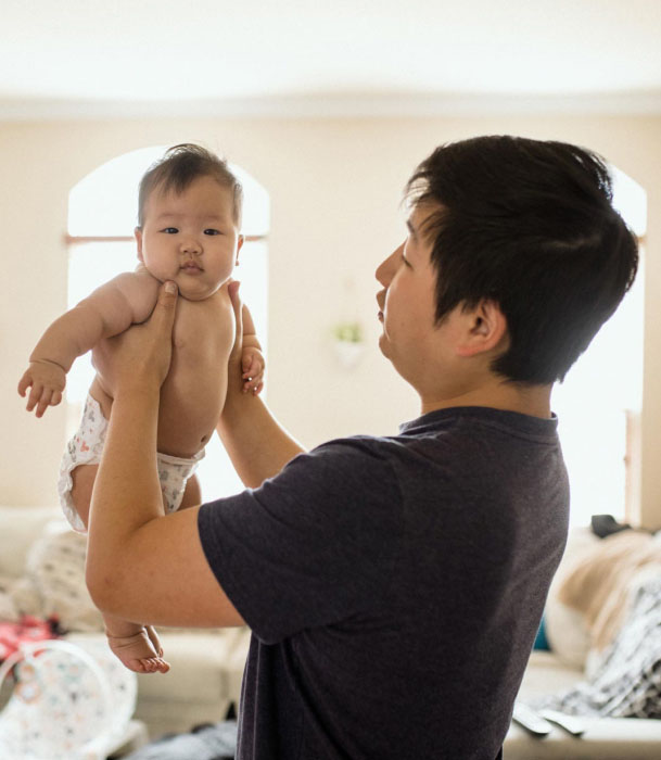 A father stands in his living room, holding his young baby up in the air.