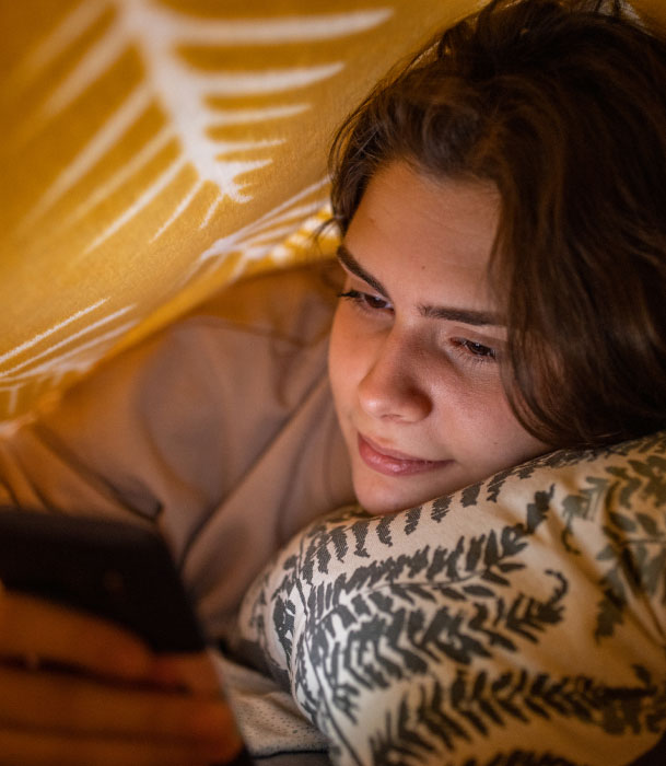 A teenage girl lying down in bed, looking at her phone under the covers.