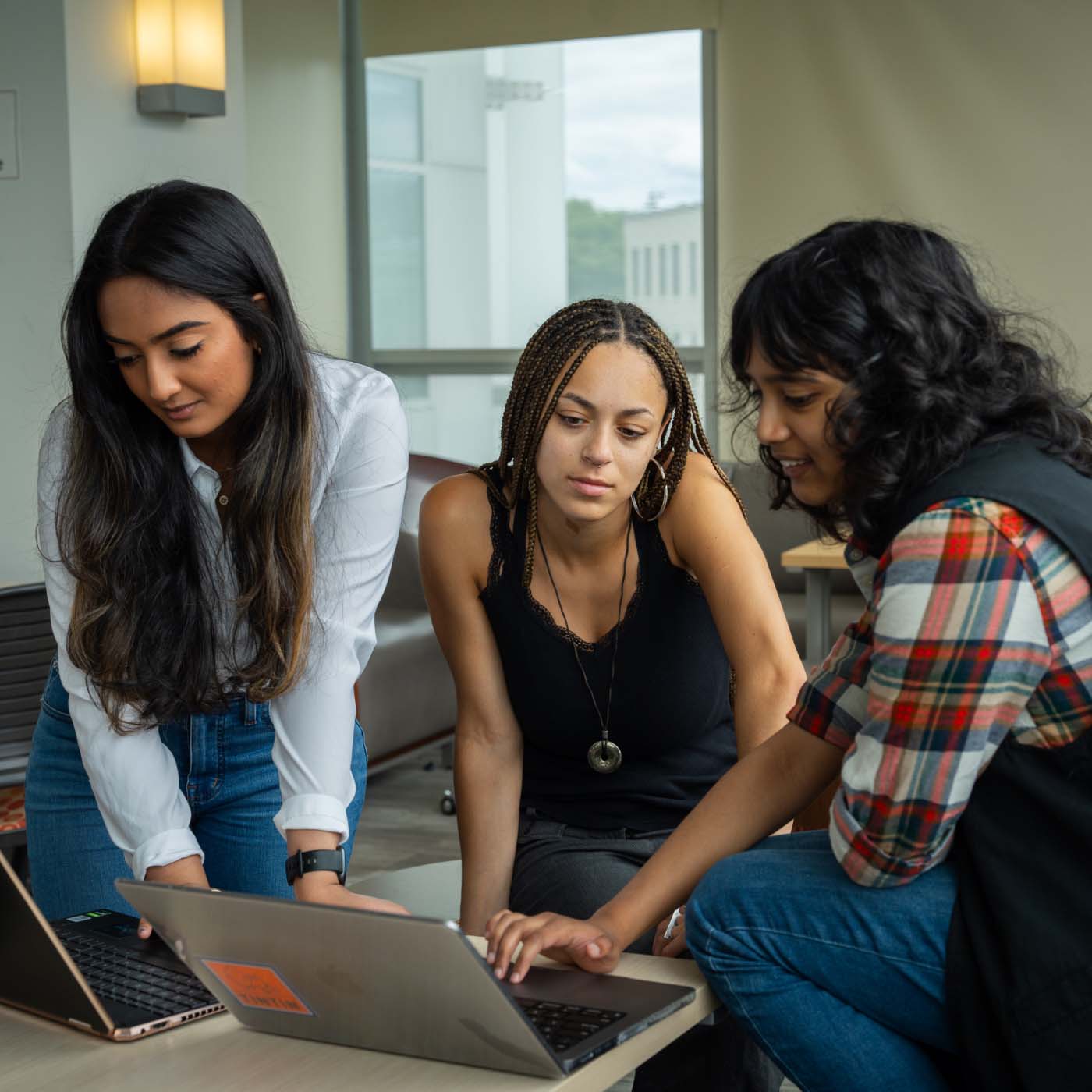 Three students talking together and looking at a single laptop at a table in a classroom