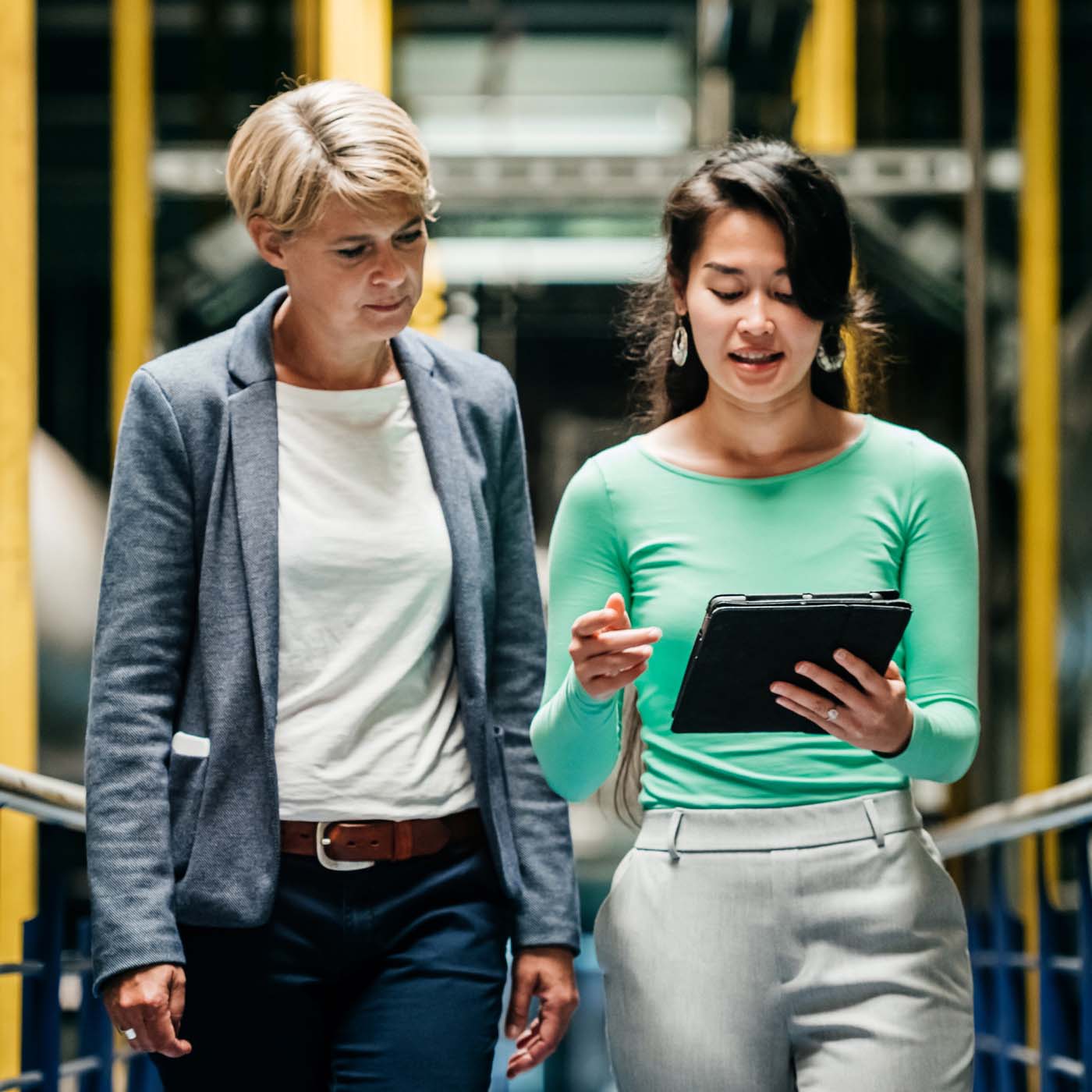 Two women talking and looking at a tablet while walking through a factory