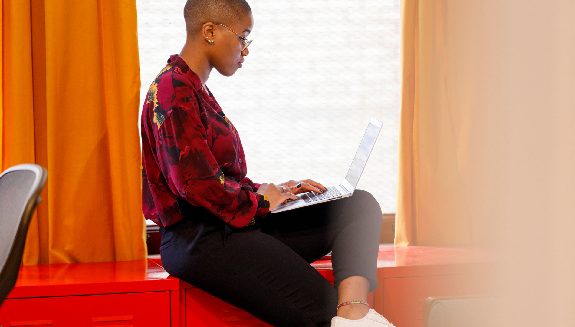 A woman working on a laptop while sitting near a windowsill in an office