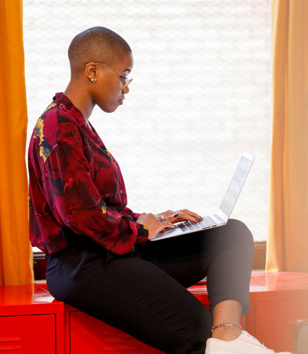 A woman working on a laptop while sitting near a windowsill in an office.